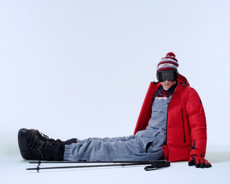 Person in ski gear wearing overalls, jacket, beanie, and goggles while sitting on a white surface with skis and poles.