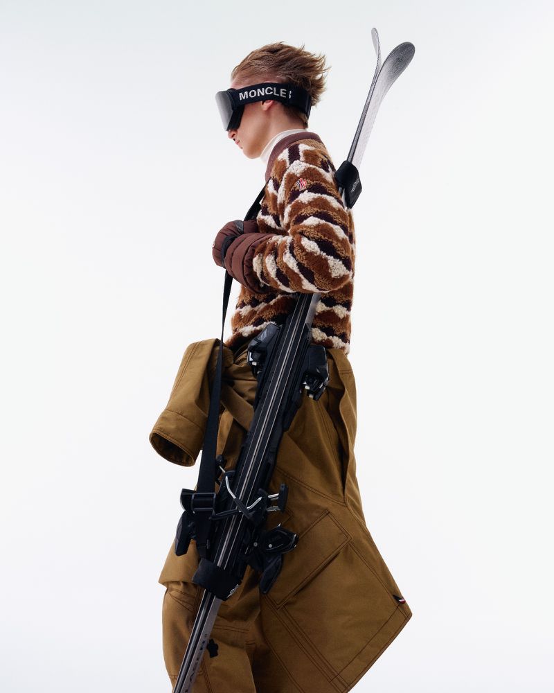 Studio shot of a person wearing ski goggles, brown patterned sweater, and tan pants, with skis and ski equipment over their shoulder.