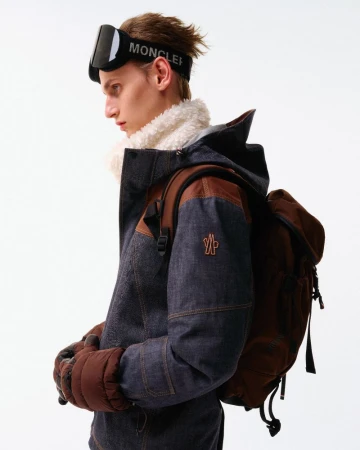 Profile shot of a man in a blue denim jacket and snow goggles, wearing a brown backpack and gloves against a white background.