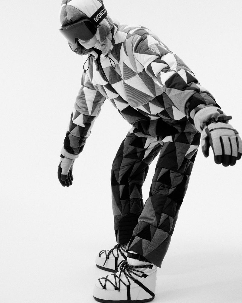 Monochrome studio shot of an adult wearing a Moncler logo visor, a quilted geometric-patterned suit, gloves, and moon boots against white.