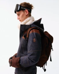 Model wearing Moncler ski goggles, puffy white collar, denim jacket, brown backpack and gloves against a white background