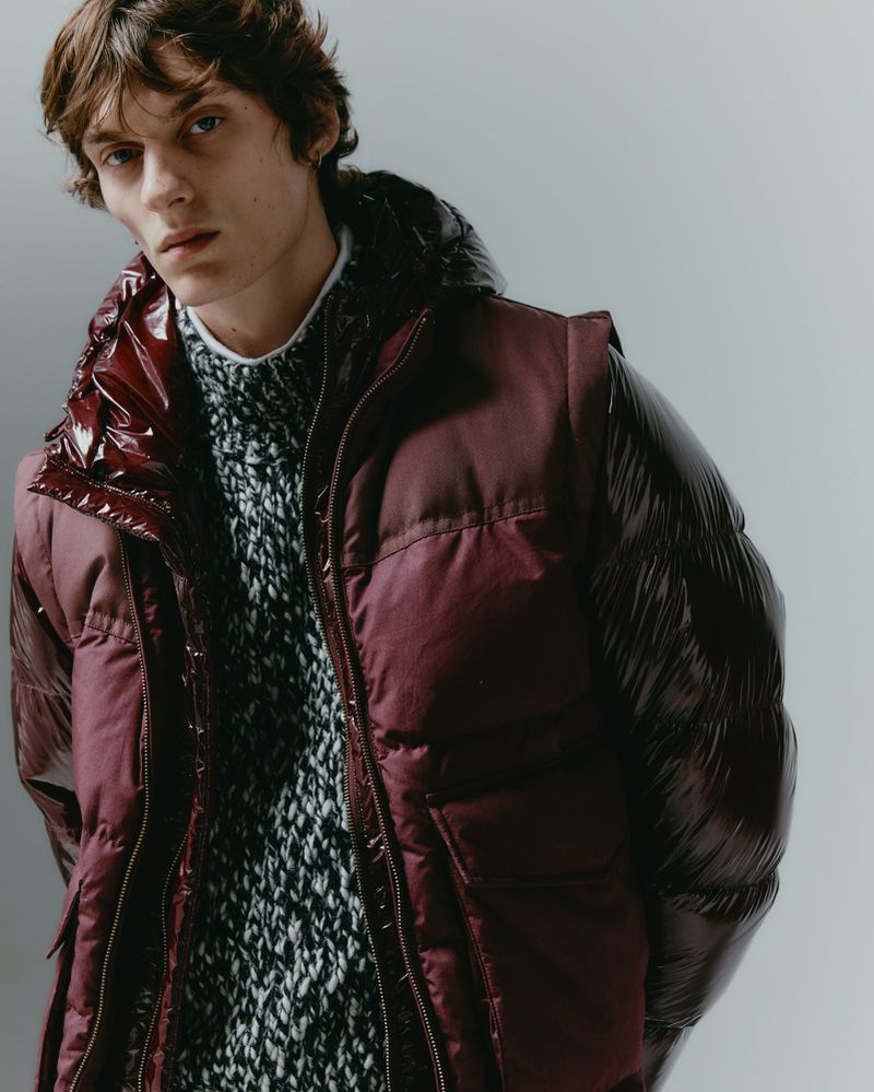 Close-up studio shot of a male model wearing a burgundy puffer vest over a grey and black patterned sweater, looking at the camera.