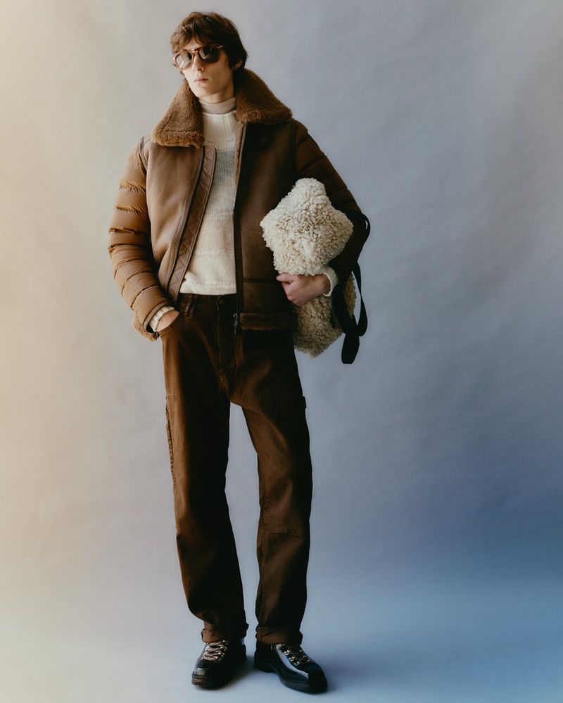 Man in brown shearling jacket and brown trousers holds a white tote bag indoors, fashion style photography, neutral background, eye-level view.