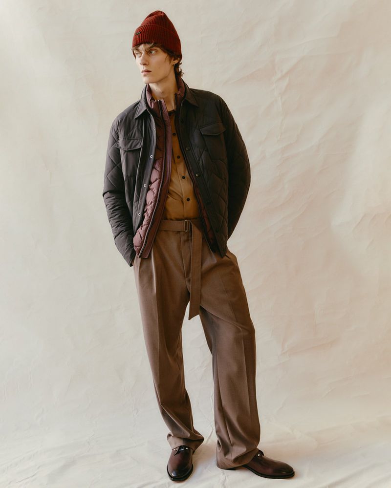 Male fashion model in a quilted jacket, rust colored pants and a beanie poses in a studio setting with a crumpled paper background.