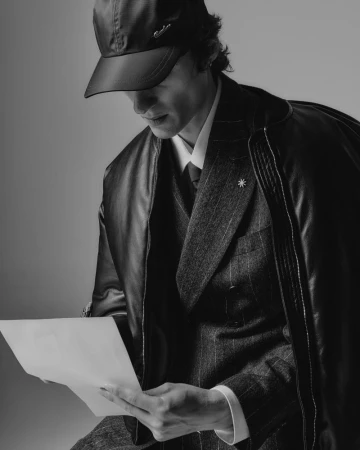 Black-and-white portrait of an adult man wearing a suit and cap, looking down at a piece of paper he holds in both hands in a studio.