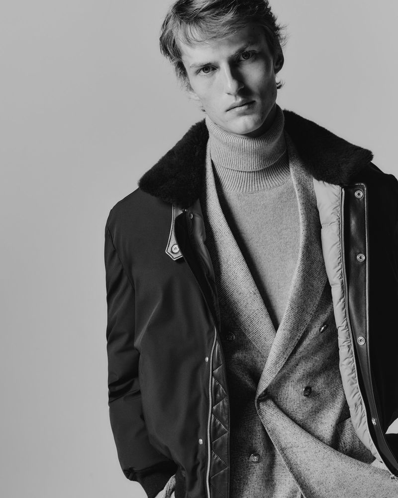 Monochrome fashion shot of a blond male model wearing a gray turtleneck sweater and a black bomber jacket with fur collar against a grey background.