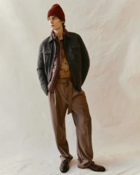 A male model wearing a quilted jacket, brown trousers, and a burgundy beanie standing against a creased backdrop.