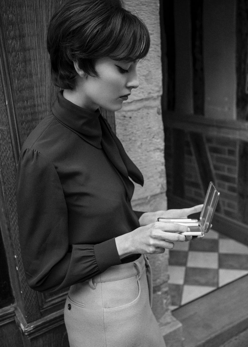 Monochrome portrait of a stylish woman with short hair in vintage fashion, holding a compact in an indoor setting with muted tones.