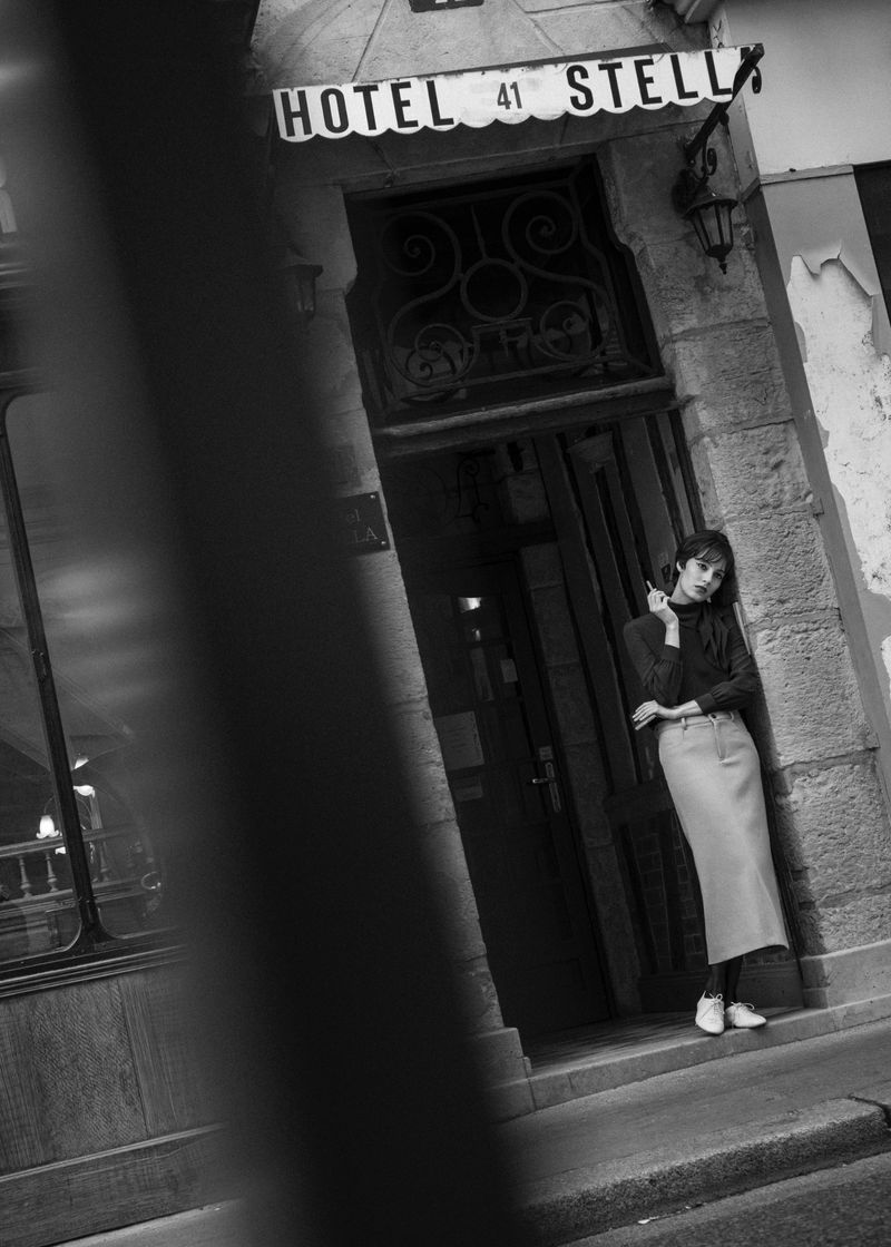 A monochrome shot of an adult female standing in the doorway of Hotel 41 Stella, wearing a long skirt, top, and sneakers.