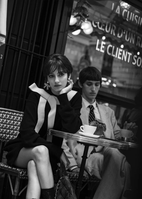 Black and white portrait of a stylish adult woman sitting at a cafe table with a man in the background, city scene.