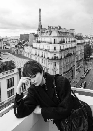 Model leaning on a balcony railing in Paris with the Eiffel Tower in the background, wearing a dark coat and shoulder bag.