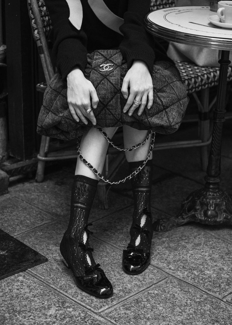 Monochrome fashion shot of a seated person with a quilted handbag and patent leather shoes on a tiled floor in an indoor setting.