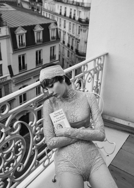 Monochrome fashion shot of a woman in lace lingerie, sitting on a balcony with an open book, overlooking city buildings.