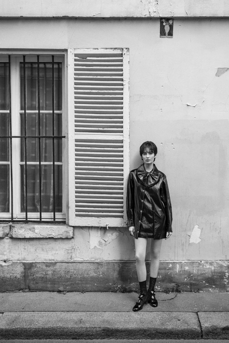 Monochrome shot of a young woman in a black coat, posing outdoors next to a weathered building with a window and shutter.