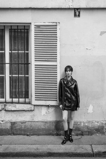 Monochrome shot of a young woman in a black coat, posing outdoors next to a weathered building with a window and shutter.