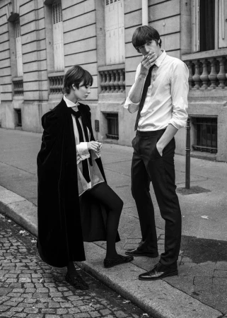 Black and white editorial image of a woman in a velvet cape and a man smoking on a city street