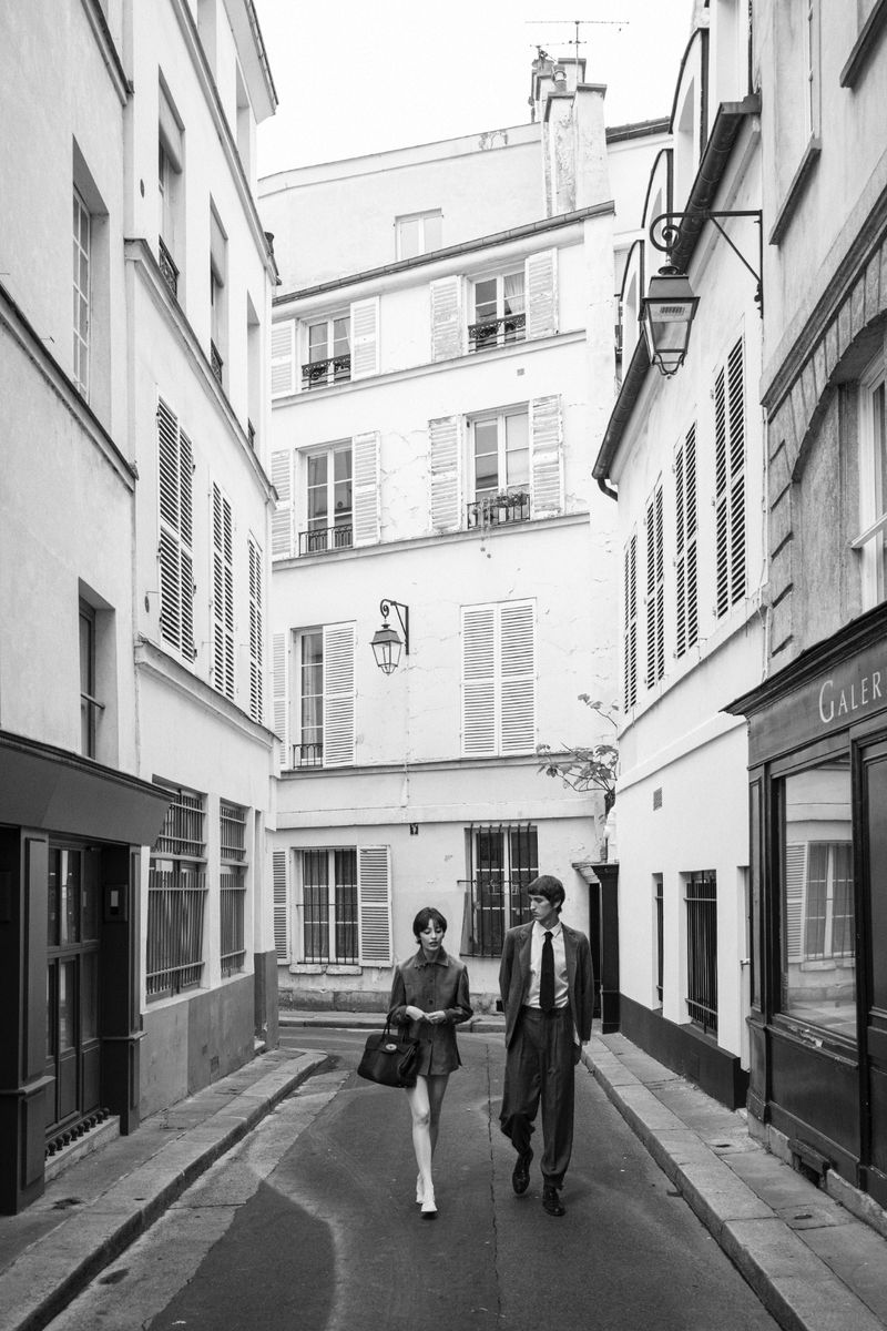 A woman and a man stroll down a narrow street lined with buildings with shutters in a black and white street scene.