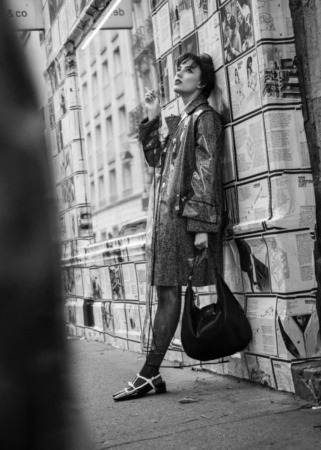 Black and white fashion shot of an adult female in a translucent coat, smoking and leaning against a newspaper-covered wall in a city.