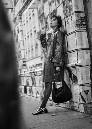 Black and white fashion shot of an adult female in a translucent coat, smoking and leaning against a newspaper-covered wall in a city.