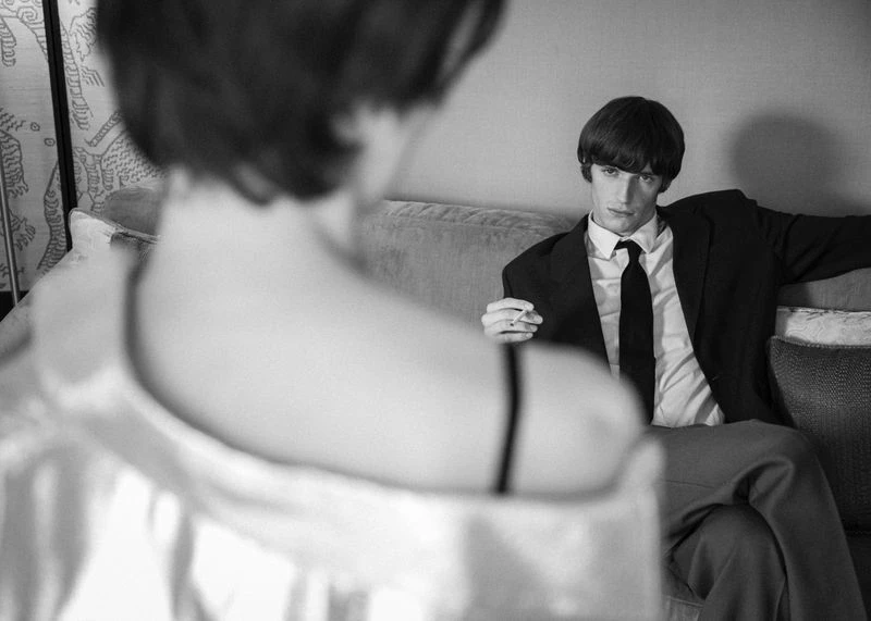 Black and white medium shot of an elegant man in a suit and tie holding a cigarette, viewed over a woman's shoulder.