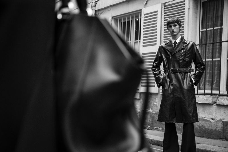 Monochrome portrait of a man in a black leather coat standing in front of a building with shuttered windows, fashion photography.