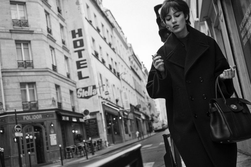 A stylish woman in a black coat stands on a Paris street, holding a purse, with classic buildings and a hotel in the background.