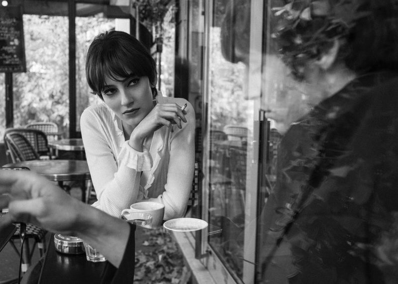 Black and white shot of a stylish woman smoking in a cafe with a serious expression, seated at a table with coffee cups.