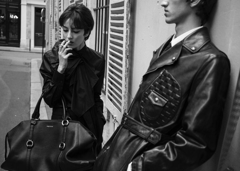 Black and white portrait of woman with cigarette, standing next to man in leather jacket on urban street with handbag.