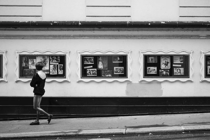 Black and white image of an adult female walking past a white building with framed vintage movie posters on display.