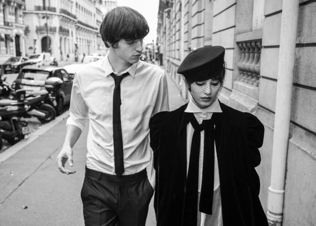 Black and white editorial image of a man in a white shirt and tie walking alongside a woman wearing a beret and dark coat on a street.
