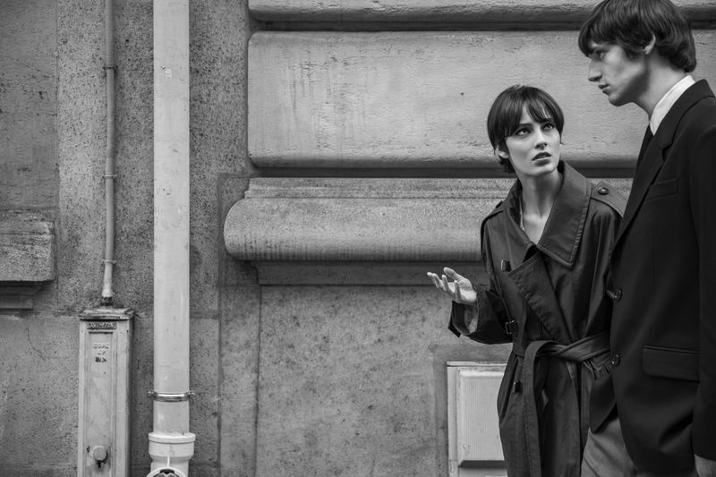 Monochrome fashion shot of a stylish adult male and female couple in coats standing next to a stone urban building in daylight.