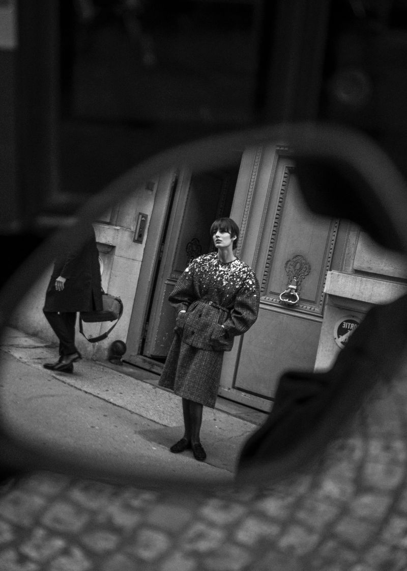 Black and white fashion shot in street mirror reflection: adult woman in patterned jacket and skirt suit with passerby in background.