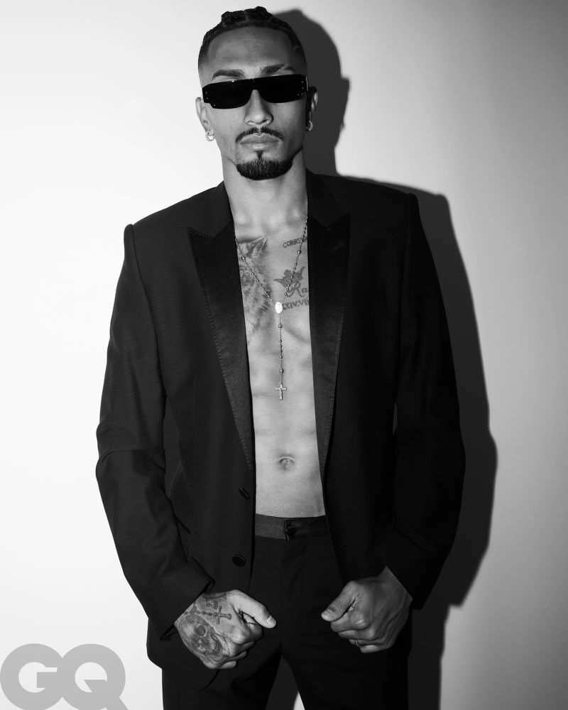 Portrait of Raphinha wearing a black tuxedo jacket with sunglasses and necklaces against a plain white background for GQ Espana.