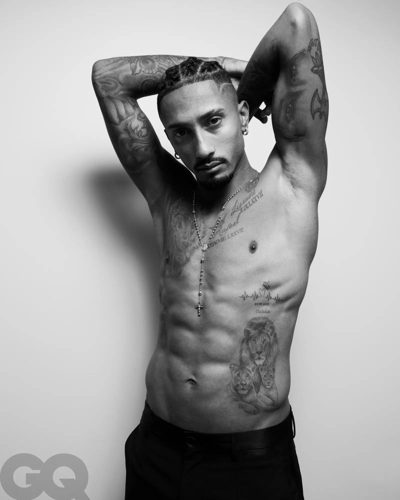 Portrait of Raphinha with arms raised, showcasing torso tattoos against a plain studio background for GQ Espana