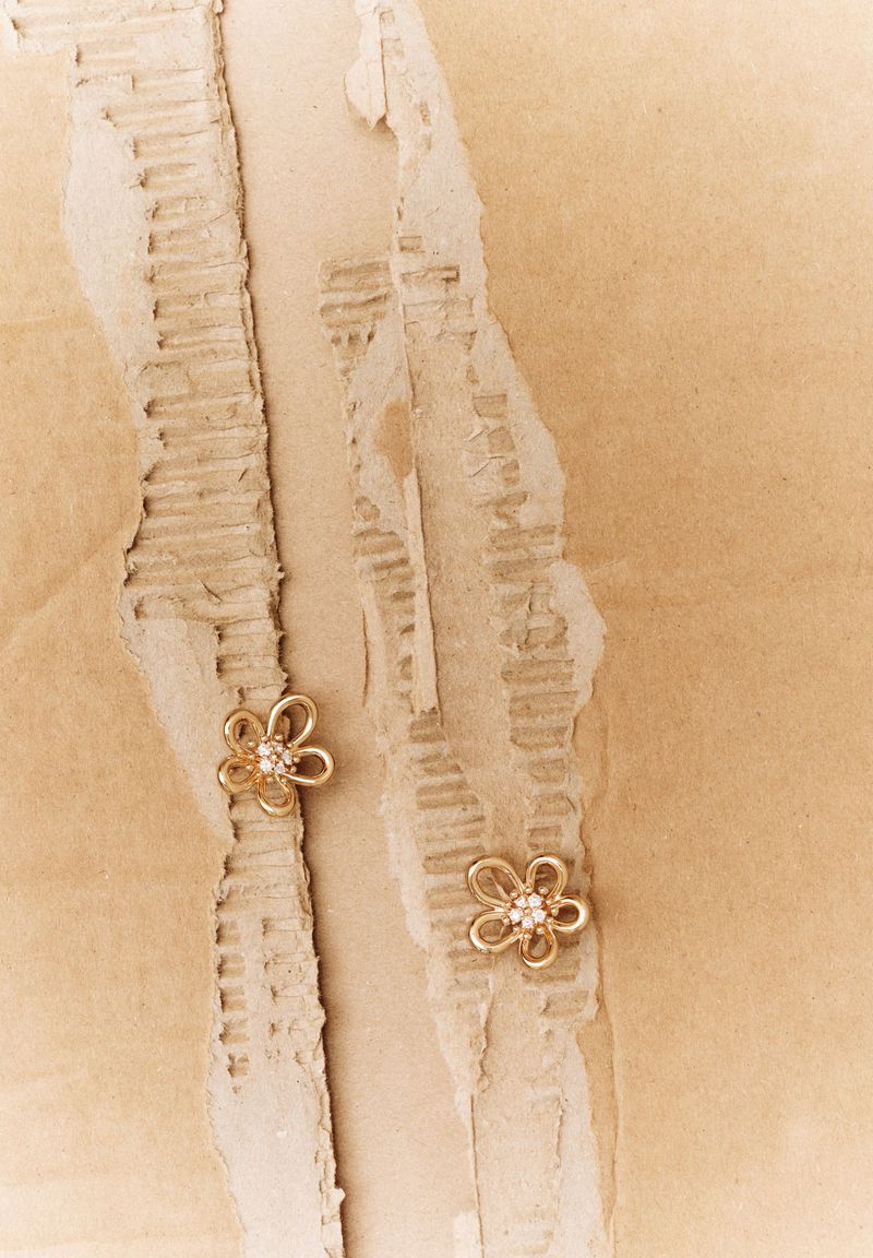 Two gold flower-shaped earrings with diamond accents on torn corrugated cardboard, warm tones, product photography, neutral background.