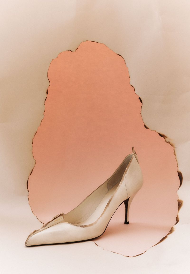 Cream-colored high-heeled shoe placed in front of a torn, irregularly-shaped pink paper backdrop on a cream surface.