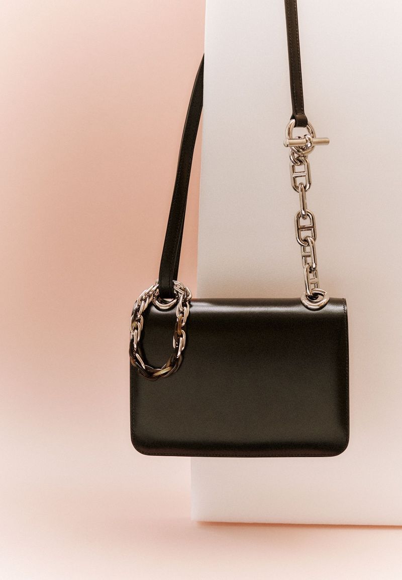 Studio shot of a black leather purse with silver chain detailing, hanging against a split pink and white background.