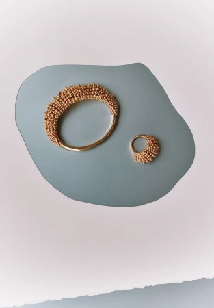 Still life of a gold bracelet and ring, both featuring a textured, beaded design, arranged on a light blue and white backdrop.