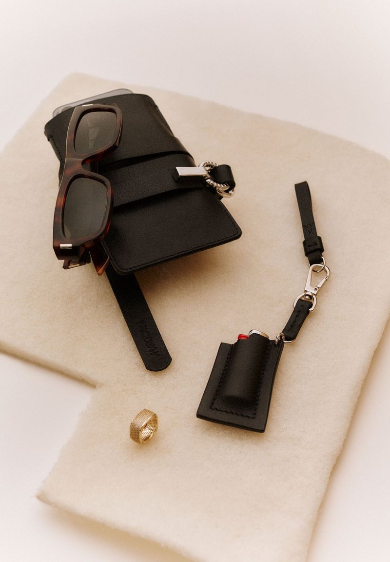 Still life studio shot featuring black leather accessories, tortoiseshell sunglasses, and a gold ring on a soft cream-colored fabric.