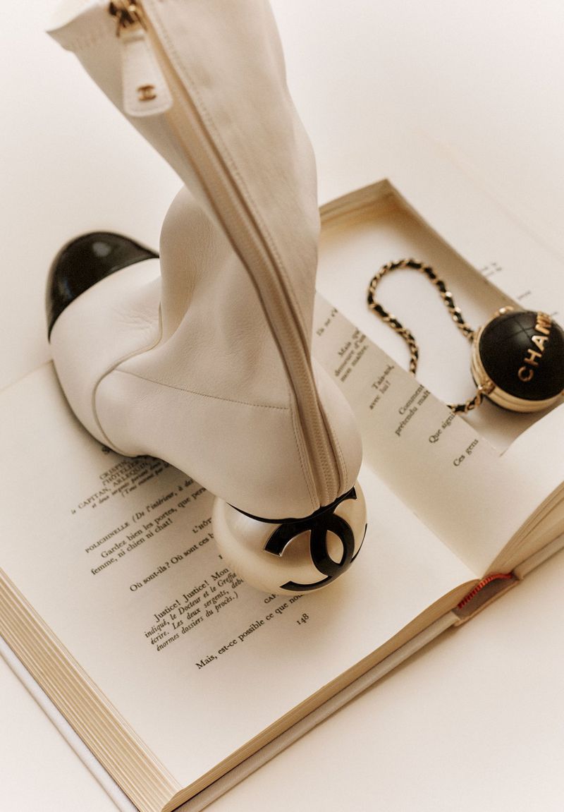 White ankle boot with a black cap toe standing upright on an open vintage book next to a small round chained bag.