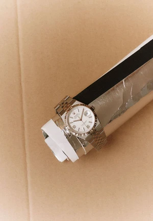 Silver luxury watch with a white dial resting on a rolled cardboard tube against a plain brown background