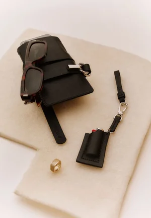 Black leather accessories, tortoiseshell sunglasses, and a ring arranged on a neutral fabric surface