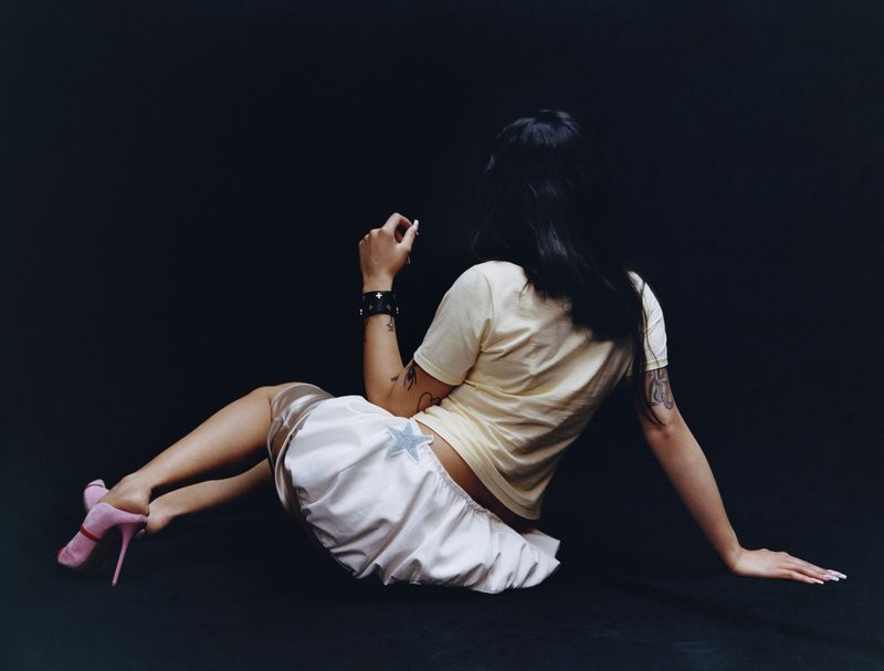 Young adult woman with black hair poses sitting on the floor in a light yellow t-shirt, satin skirt with a star, and pink heels.
