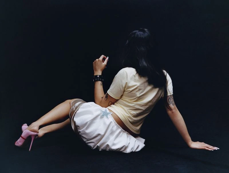 Young adult woman with black hair poses sitting on the floor in a light yellow t-shirt, satin skirt with a star, and pink heels.