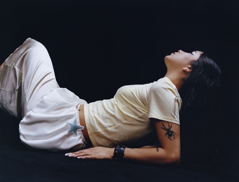 Young woman with dark hair, wearing white pants and a pale yellow shirt, poses against a dark background, looking up.
