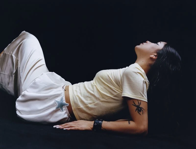 Young woman with dark hair, wearing white pants and a pale yellow shirt, poses against a dark background, looking up.