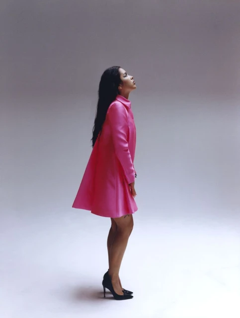 Woman in pink dress and black high heels poses in studio. She's looking upward in a fashion pose. The studio has a neutral background.