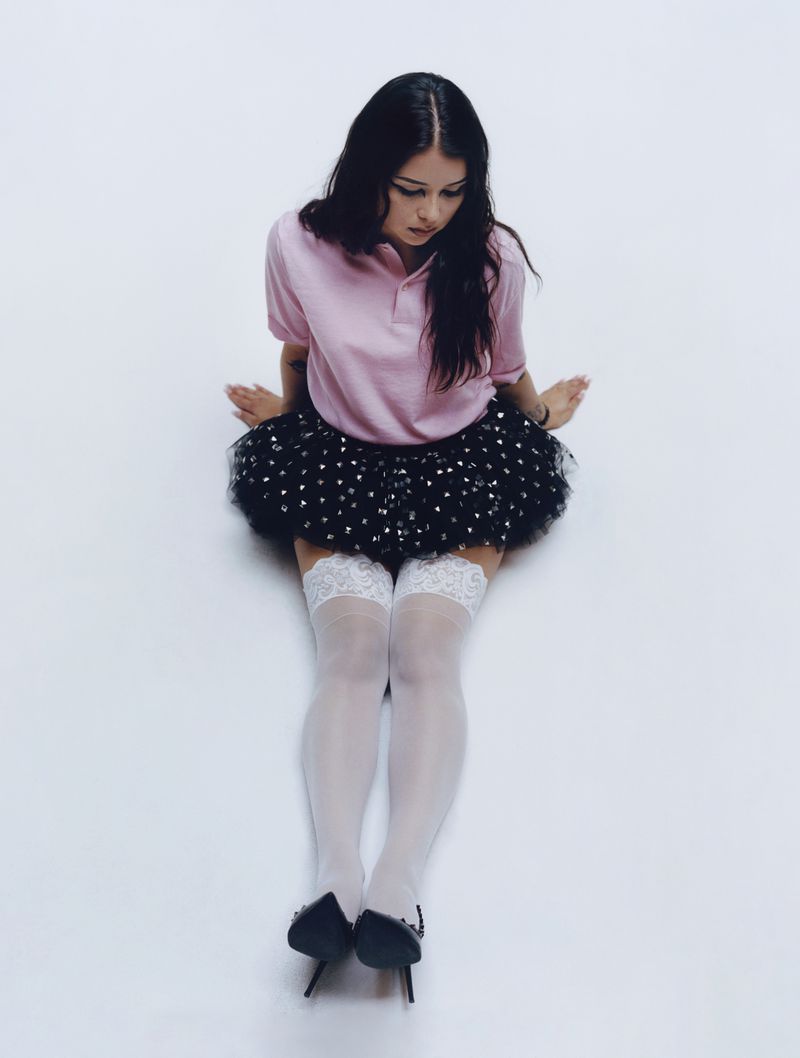 Young woman in pink polo shirt and black tutu-style skirt poses in studio. Sheer white stockings and black heels complete the fashion look.