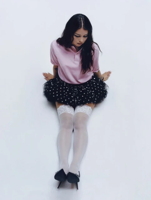 Young woman in pink polo shirt and black tutu-style skirt poses in studio. Sheer white stockings and black heels complete the fashion look.