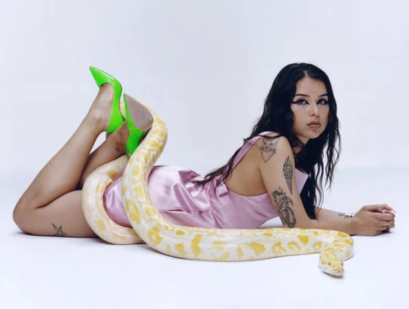Woman in pink dress lying down with yellow snake and green heels in white studio. Fashionable beauty portrait in studio.